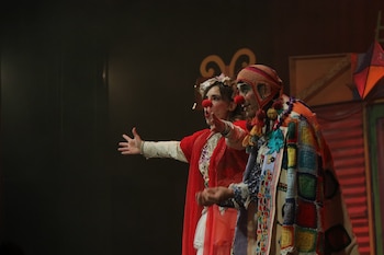 Dos actores con narices de payaso, vestuario colorido y maquillaje actúan en un escenario oscuro con elementos decorativos al fondo