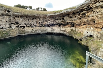 Pozo circular con agua verde oscura, paredes de roca erosionada y agujeros, vegetación seca en bordes superiores, barandilla metálica, objeto rojo en el agua