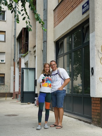 Un hombre con mochila y gafas y una mujer con auriculares posan sonrientes en una acera junto a un edificio