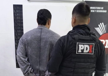Un hombre de espaldas en sudadera gris parado frente a una tabla de medición de altura, observado por un oficial de PDI con chaleco táctico