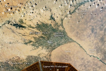 Fotografía satelital con terreno desértico, zonas verdes rotuladas y el panel solar de una nave espacial en el borde inferior