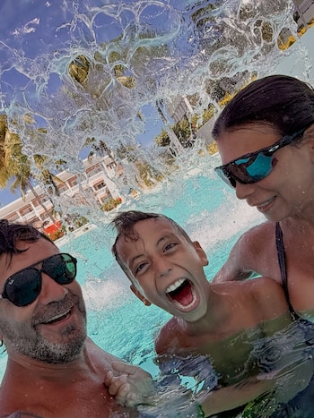 Un hombre con gafas oscuras, un niño sonriente y una mujer posan dentro de una piscina detrás de una cortina de agua