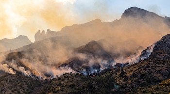 Laderas montañosas rocosas cubiertas por humo espeso con varios focos de fuego visibles en la superficie.