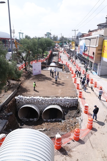 Vista aérea de un sitio de construcción con excavaciones, tubos metálicos de drenaje, sacos de arena, conos de tráfico, operarios y peatones en una calle