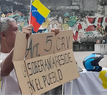 Un hombre de cabello gris y camisa blanca sujeta una pancarta de cartón y una pequeña bandera de Venezuela. Al fondo, una pared con grafitis y un casco azul