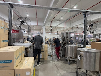 Vista general del interior de una fábrica con operarios, máquinas de llenado y etiquetado de botellas, y cajas de "Piroplast" apiladas. Líneas de producción visibles