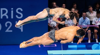 Dos clavadistas masculinos en el aire sobre una piscina azul, una plataforma blanca, y una pared azul con letras blancas, público de fondo.