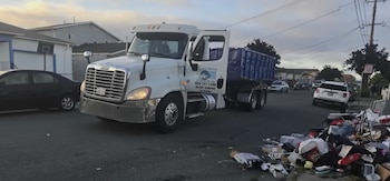 Un camión blanco de Express Rental con un contenedor de basura azul en la parte trasera se ve en una calle residencial junto a un montón de basura en la acera