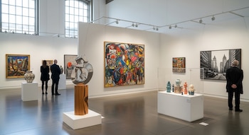 Galería de arte moderna con paredes blancas, ventanales y suelo brillante. Se aprecian pinturas, esculturas y vasijas. Varias personas observan las obras expuestas.