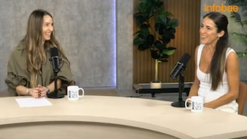 Dos mujeres conversan en un estudio frente a micrófonos y tazas sobre un escritorio curvo de madera clara