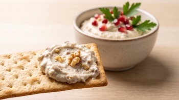 Galleta salada con dip de crema y nuez frente a un recipiente con crema, granos de granada y perejil sobre una mesa.