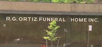 Letrero dorado de R.G. Ortiz Funeral Home Inc. sobre fachada de piedra oscura con un pequeño árbol al frente