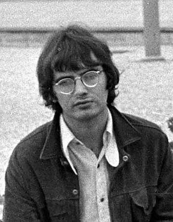 Retrato en blanco y negro de Tom McGuinness, un hombre con cabello oscuro de longitud media y gafas redondas, vistiendo una camisa de cuello y una chaqueta oscura