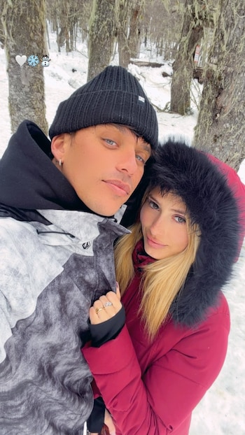 Un hombre con gorro negro y abrigo estampado posa junto a una mujer con abrigo rojo con capucha de piel en un bosque con nieve