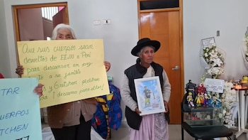 Dos mujeres sostienen carteles y una fotografía enmarcada junto a arreglos florales y figuras de acción sobre una mesa en una habitación