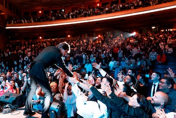 Dante Gebel en traje oscuro se inclina hacia una multitud que levanta las manos en un auditorio abarrotado, bajo luces brillantes