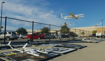 Drones de entrega blancos con detalles amarillos en el aire y en el suelo en un área de prueba. Una valla, coches y un edificio comercial son visibles al fondo bajo un cielo azul