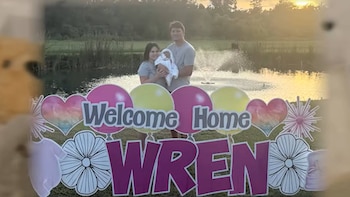Un hombre y una mujer sostienen a una bebé en un jardín frente a carteles decorativos con el texto Welcome Home y WREN