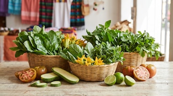 Canastas de mimbre repletas de quelites frescos, hojas verdes y flores comestibles, con jitomates, calabacitas y limas cortadas sobre una mesa de madera rústica.