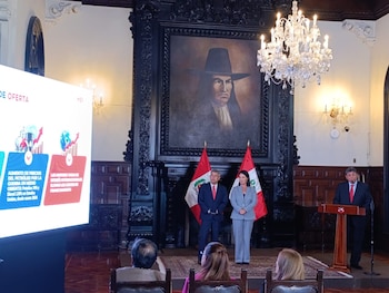 Tres funcionarios vestidos con traje formal participan en una conferencia frente a dos banderas peruanas, una pantalla y una pintura histórica