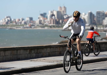 Hombre en bicicleta pedalea por la calle costera, bebe de una botella. Viste casco, lentes, camiseta blanca y shorts. Detrás, el mar y una ciudad