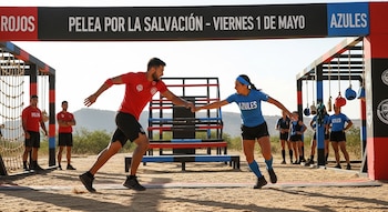 Atleta de rojo y atleta de azul se tocan las manos en pista de arena. Al fondo, compañeros y estructura de obstáculo bajo cielo.