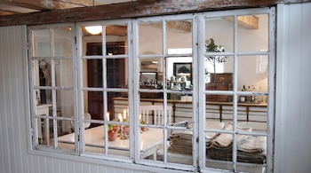 Una ventana de madera blanca con múltiples paneles ofrece una vista de un comedor rústico con una mesa, sillas, velas encendidas y plantas en macetas.