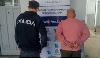 Vista trasera de un oficial de policía y un hombre calvo con suéter rosa, frente a un cartel de la Policía de Seguridad La Plata