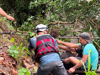 Tres rescatistas con uniformes de la Cruz Roja, cascos y chalecos, junto a un vehículo de rescate blanco. El fondo muestra un paisaje montañoso y húmedo con vegetación