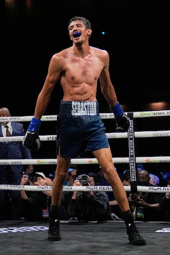 Un boxeador de pie en un cuadrilátero lleva pantalones cortos oscuros, guantes y un protector bucal, rodeado por fotógrafos tras las cuerdas