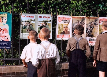 Vista trasera de cuatro niños mirando carteles de propaganda colgados en una valla metálica. Los carteles muestran imágenes de guerra y figuras políticas