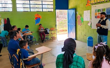 Un hombre barbudo habla a un grupo de adultos y niños sentados en pupitres de un aula con paredes verdes, ventanas y una puerta abierta al exterior