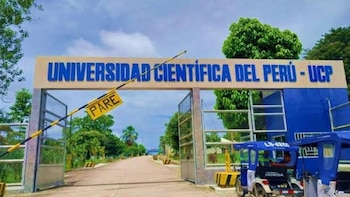 Entrada principal de la Universidad Científica del Perú con un cartel identificativo, una barrera de seguridad que dice "PARE" y motocarros estacionados