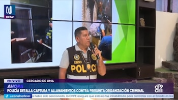 Un oficial de policía peruano con chaleco "POLICÍA" habla en un micrófono frente a pantallas de televisión que muestran imágenes de allanamientos policiales y un logo de noticias