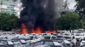 Vista de un gran incendio con llamas intensas y humo negro sobre un estacionamiento lleno de coches, con edificios y árboles al fondo