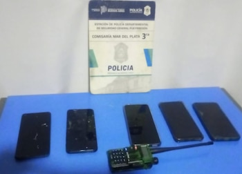 Placa de la Comisaría Mar del Plata 3ra, cinco teléfonos celulares negros y un walkie-talkie verde con antena negra, sobre una superficie azul