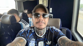 Hombre con gorra, gafas de sol y camiseta de Argentina, brazos y cuello tatuados, toma selfie dentro de un autobús con otros pasajeros detrás.