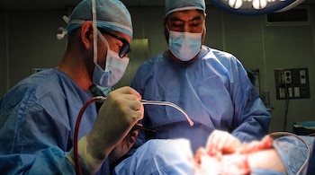 Dos cirujanos vestidos con batas, mascarillas y gorros quirúrgicos azules manipulan instrumentos médicos durante una operación