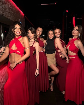Seis mujeres jóvenes sonríen y posan en un interior con iluminación rojiza; cinco visten vestidos rojos o borgoña, y una lleva un top negro y falda beige