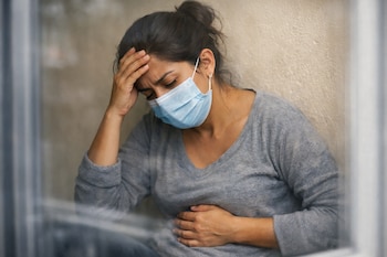 Mujer latina con mascarilla, una mano en la cabeza y la otra en el estómago, mira hacia abajo, delante de una pared beige y un vidrio.