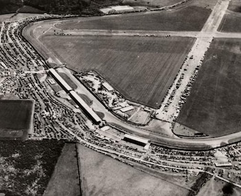 Fotografía aérea en blanco y negro de un circuito de carreras con pistas, edificios, gradas, zonas de estacionamiento llenas de vehículos y campos