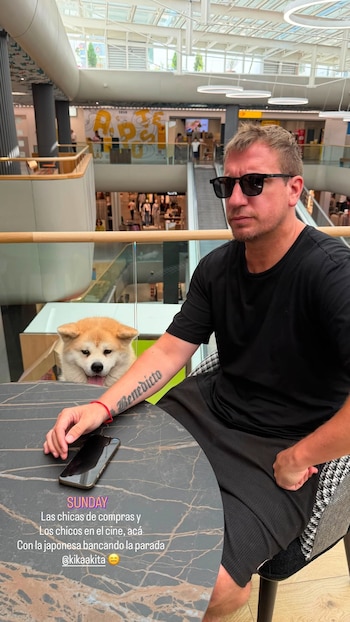 Hombre con gafas de sol y camiseta negra sentado en una mesa. A su lado, un perro Akita de pelaje claro. En la mesa hay un teléfono. Fondo de centro comercial