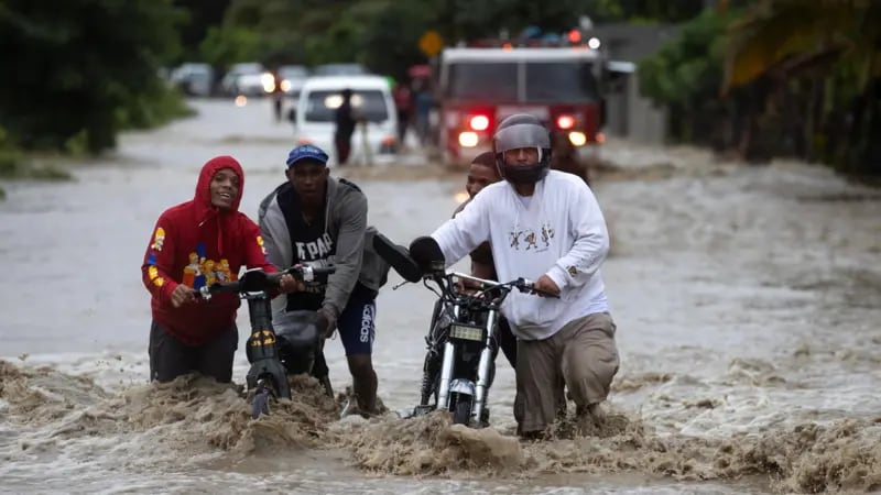 Varias personas avanzan con dificultad por una calle inundada, empujando motocicletas tras las intensas lluvias registradas en República Dominicana (Foto cortesía BBC).