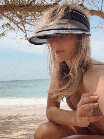 Mujer de cabello rubio con visor negro en una playa de arena clara, el mar azul con boyas amarillas y un árbol que le da sombra