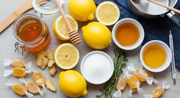 Primer plano de ingredientes para caramelos: limones enteros y cortados, frascos de miel, jengibre, azúcar en un cuenco y caramelos envueltos sobre madera clara.