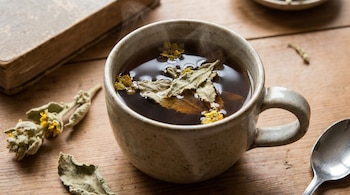 Primer plano de una taza de té de gordolobo humeante con hojas y pequeñas flores amarillas flotando, junto a un libro antiguo y una cuchara sobre madera.