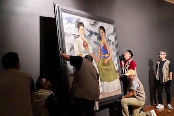 Cinco hombres instalan un cuadro grande con marco gris de una obra tipo Las Dos Fridas en una pared gris oscura de una galería
