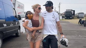 Nicole Neumann, con gafas y chaleco, sostiene a una niña con protectores. Camina junto a un hombre con gorra y traje de piloto en un autódromo con vehículos
