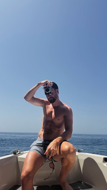 Un hombre sin camisa y con bañador en un barco. Sostiene una cámara fotográfica cerca de su rostro y gafas de sol en la otra mano. Mar y cielo azul