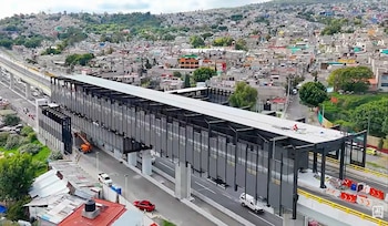 Estación elevada con estructura metálica gris sobre columnas de concreto, sobre una avenida con automóviles y casas distribuidas en una colina